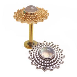 Peut inclure: Deux bijoux : un piercing pour la lèvre doré et un piercing pour la lèvre argenté. Chaque bijou présente une pierre précieuse ronde et irisée sertie dans un cadre décoratif en forme d'éclat de soleil. Le bijou doré est droit, tandis que le bijou argenté est à plat.