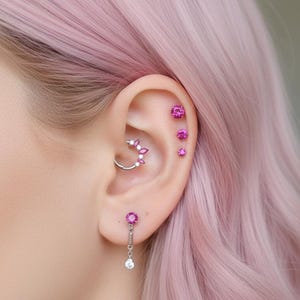 May include: Close-up of an ear adorned with multiple piercings. The ear features a silver hoop earring with pink gemstones, along with several pink gem studs. The person has pink hair, complementing the jewelry.