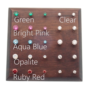 May include: A wooden display board with rows of small, round, colorful gemstones. The gemstones are labeled "Green", "Bright Pink", "Aqua Blue", "Opalite", and "Ruby Red". There are also rows of clear gemstones labeled "Clear".
