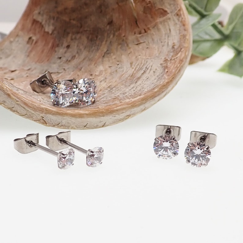 May include: A set of three pairs of silver stud earrings with clear cubic zirconia stones. The earrings are displayed on a wooden surface.