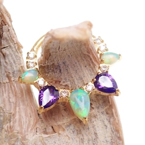 May include: A gold ring with a series of gemstones. The ring features pear-shaped opals in green and white, amethyst stones, and small clear crystals. The gemstones are set in a circular pattern. The ring is displayed on a wooden surface.