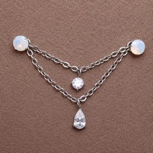 Könnte beinhalten: Ein silberfarbenes Körperschmuckstück mit zwei runden, schillernden weißen Cabochon-Steinen, die durch zwei drapierte Metallketten verbunden sind. Ein kleiner, runder, klar facettierter Stein hängt an der oberen Kette und ein birnenförmiger Stein unten.