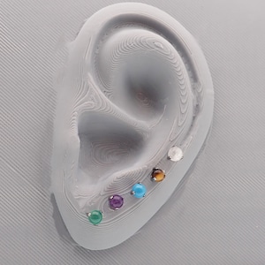 May include: A close-up of a model ear with six different gemstone earrings. The earrings are in a line, starting with a white stone, then a brown tiger's eye, a turquoise stone, a purple amethyst, a green stone, and a dark green stone.