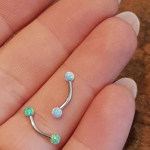 May include: Two curved barbell body jewelry pieces with metallic shafts. One piece features two light blue opal-style spheres, while the other piece features two bright green opal-style spheres at each end of the curved metal bar.
