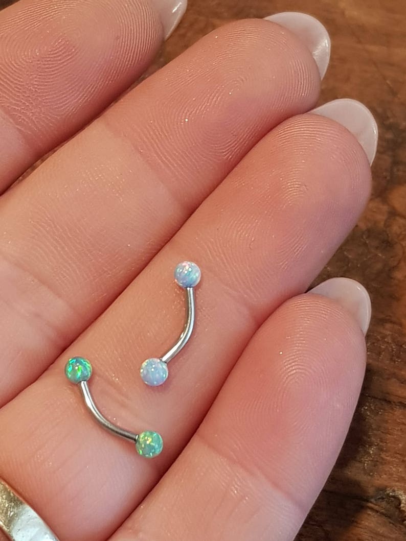 May include: Two curved barbell body jewelry pieces with metallic shafts. One piece features two light blue opal-style spheres, while the other piece features two bright green opal-style spheres at each end of the curved metal bar.