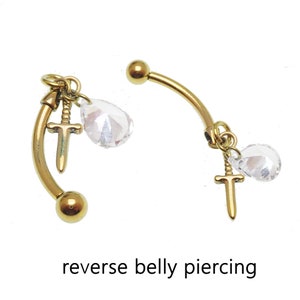 May include: A pair of gold-toned belly button rings with a dangling sword charm and a clear crystal teardrop. The rings are curved and have a ball at the end. The text "reverse belly piercing" is written below the rings.