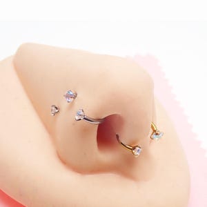 May include: A selection of nose piercings, including a curved barbell and a hoop, each featuring a clear, iridescent gemstone. Several small, stud-style nose rings with similar gemstones are also visible. The jewellery is set against a light skin tone.