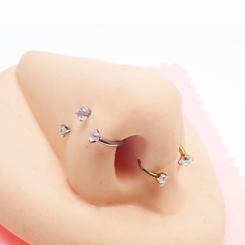 May include: A selection of nose piercings, including a curved barbell and a hoop, each featuring a clear, iridescent gemstone. Several small, stud-style nose rings with similar gemstones are also visible. The jewellery is set against a light skin tone.