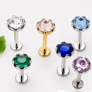 May include: A collection of six labret body jewelry pieces featuring round faceted gemstones in clear, light pink, light blue, dark blue, green, and purple. Each stud has a flat circular base and a prong setting with a scalloped metal edge.