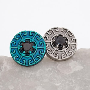 May include: Two circular buttons featuring a Greek key pattern border and a central black faceted stone. One button has a metallic teal finish, while the other has a metallic silver finish. Both items are round with raised edges and intricate geometric detailing.