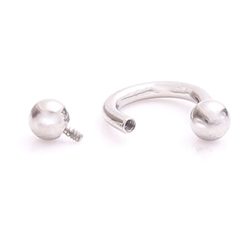 Pu&ograve; includere: Un piercing a forma di ferro di cavallo argentato con una sfera staccabile. Il bilanciere curvo ha un'estremit&agrave; filettata per avvitare la sfera. La sfera &egrave; staccata, mostrando la vite. Il gioiello &egrave; in metallo lucido e lucido.
