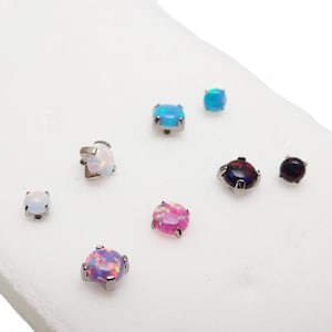 May include: A collection of opal stud earrings in various colors, including blue, pink, purple, and black. The earrings have a round or square shape and are set in silver-colored metal. The opals have a shimmering, iridescent quality.