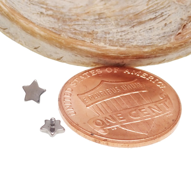 May include: Two small silver-toned star-shaped earring studs, one showing the front face and the other showing the back with a threaded post, positioned next to a copper United States one cent coin for scale to demonstrate the size of the jewelry.