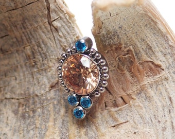 May include: A silver body jewelry piece with a round, peach-colored gemstone surrounded by smaller, clear gemstones. The piece is accented with four blue gemstones.