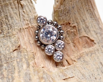 May include: A silver body jewelry piece with a cluster of clear cubic zirconia stones. The piece is designed with a large central stone surrounded by smaller stones.