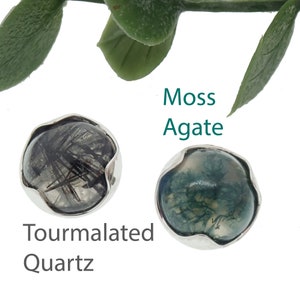 May include: Two gemstone cabochons with silver-colored settings. One is tourmalated quartz with black inclusions, the other is moss agate with green and white patterns. The text reads "Moss Agate" and "Tourmalated Quartz".