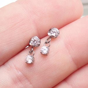 May include: A pair of silver-toned drop earrings, each featuring two clear, round-cut faceted stones. The top stone is set directly into the earring base, while the second stone hangs below from a small circular metal link, creating a dangling effect.