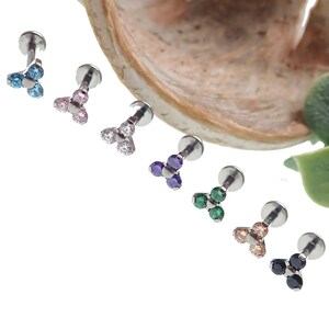 May include: A collection of seven labret studs with a three-stone design. Each stud features a different color of cubic zirconia stones, including blue, pink, clear, purple, green, peach, and black. The studs have a silver-colored post and backing.