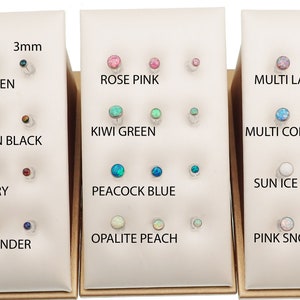 May include: A collection of round stud earrings displayed on three white stands, labeled with color names and sizes of 3mm, 4mm, and 5mm. The studs feature various iridescent colors including Galaxy Green, Harlequin Black, Dark Cherry, Sleepy Lavender, Rose Pink, Kiwi Green, Peacock Blue, Opalite Peach, Multi Lavender, Multi Cornflower Blue, Sun Ice White, and Pink Snow.