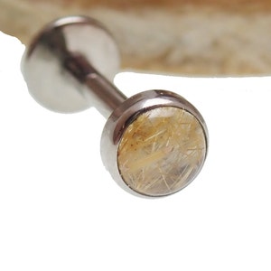 May include: A silver barbell with a round, clear, gold-toned gemstone. The gemstone has a sparkly, glittery appearance.