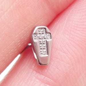 Puede incluir: Un pendiente plateado en forma de ataúd con un diseño de cruz. La cruz y la parte inferior del ataúd están adornadas con pequeñas piedras brillantes. El pendiente se muestra sobre un fondo rosa borroso.