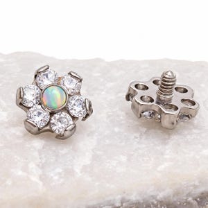 Könnte beinhalten: Ein blumenförmiges Körperschmuckstück mit einem zentralen, irisierenden Opal-Cabochon, umgeben von sechs klaren, rund geschliffenen Steinen in einer silberfarbenen Metallfassung. Die Rückseite zeigt einen Gewindestift zur sicheren Befestigung.