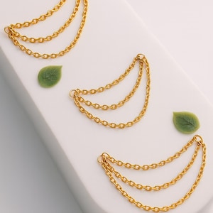 May include: Gold-coloured chain earrings, each with three strands, arranged in a curved design. The earrings are presented on a white surface, with two green leaf accents.