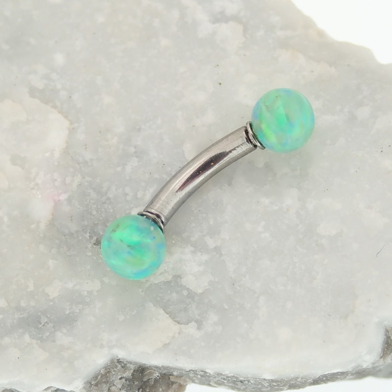 May include: A curved barbell body jewelry piece featuring a polished silver-tone metal shaft with two round, iridescent light green opal-style beads at each end. The beads display a subtle play of color with hints of blue and yellow throughout.
