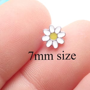 May include: A small, daisy-shaped charm with white petals and a yellow centre. The charm is approximately 7mm in size. The image shows the charm resting on a fingertip.