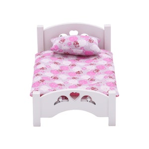 May include: A white wooden doll bed with a heart-shaped headboard and footboard. The bed is covered with a pink and white patterned blanket and pillow, featuring a repeating design of teddy bears.