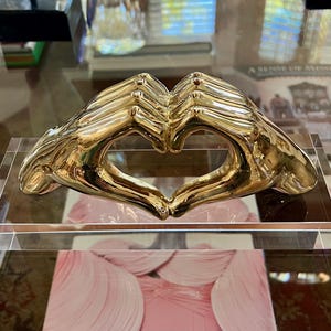 Könnte beinhalten: Eine goldfarbene Skulptur aus zwei Händen, die eine Herzform bilden. Die Skulptur ist auf einem transparenten, rechteckigen Sockel montiert. Die Hände sind detailliert und scheinen aus Metall zu sein. Die Skulptur wird auf einer Glasoberfläche ausgestellt.