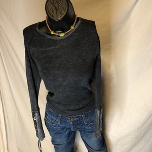 Puede incluir: Una camisa negra de manga larga con un aspecto desgastado y detalles de encaje en los puños. La camisa se combina con unos vaqueros azules.