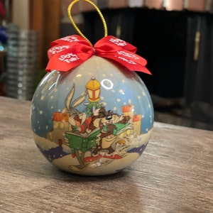 May include: A round Christmas ornament featuring Bugs Bunny and Taz the Tasmanian Devil singing carols. The ornament is decorated with a snowy winter scene and a red bow.