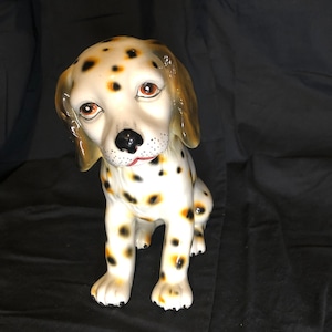 Op de afbeelding: Een keramiek beeldje van een Dalmatiër die zit met zijn hoofd gekanteld. De hond is wit met zwarte vlekken en heeft bruine oren. Het beeldje is geschilderd met realistische details, waaronder bruine ogen en een roze tong.