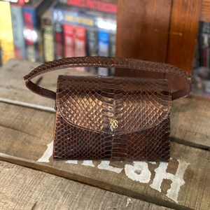Può includere: Una piccola borsa a mano in pelle marrone con motivo a pelle di serpente. La borsa ha una chiusura dorata e una tracolla corta.