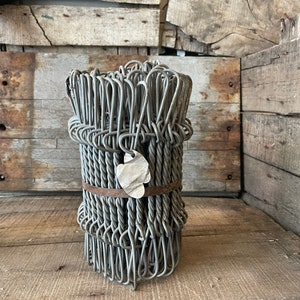 Op de afbeelding: Een grijze metalen draadvaas gemaakt van een bundel draadlussen. De vaas heeft een rustieke uitstraling en is vastgebonden met een bruine leren band.