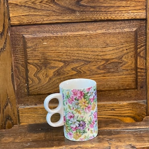 Può includere: Tazza in ceramica bianca con doppia maniglia. La tazza presenta un motivo floreale con fiori rosa, viola, gialli e verdi su sfondo bianco.
