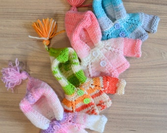 May include: A collection of handmade miniature knitted sweaters and matching hats in various colors. The sweaters feature button closures and are adorned with tassels. Colors include pink, blue, green, orange, and white.
