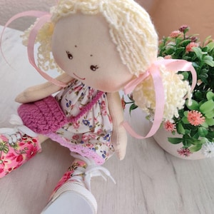 May include: A handmade doll with blonde yarn hair tied with pink ribbons. The doll wears a floral print dress, a pink crocheted purse, and floral print sneakers. A small potted plant with pink and white flowers is in the background.