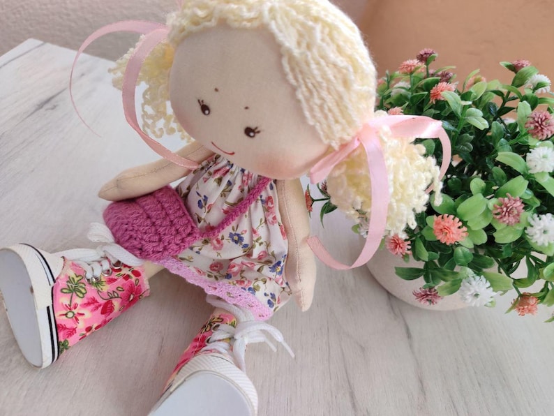 May include: A handmade doll with blonde yarn hair tied with pink ribbons. The doll wears a floral print dress, a pink crocheted purse, and floral print sneakers. A small potted plant with pink and white flowers is in the background.