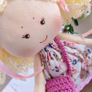 May include: A handmade cloth doll with blonde yarn hair and a painted face. The doll wears a floral print dress with a pink crocheted purse. Pink ribbons accent the doll's hair and dress, creating a whimsical and charming aesthetic.