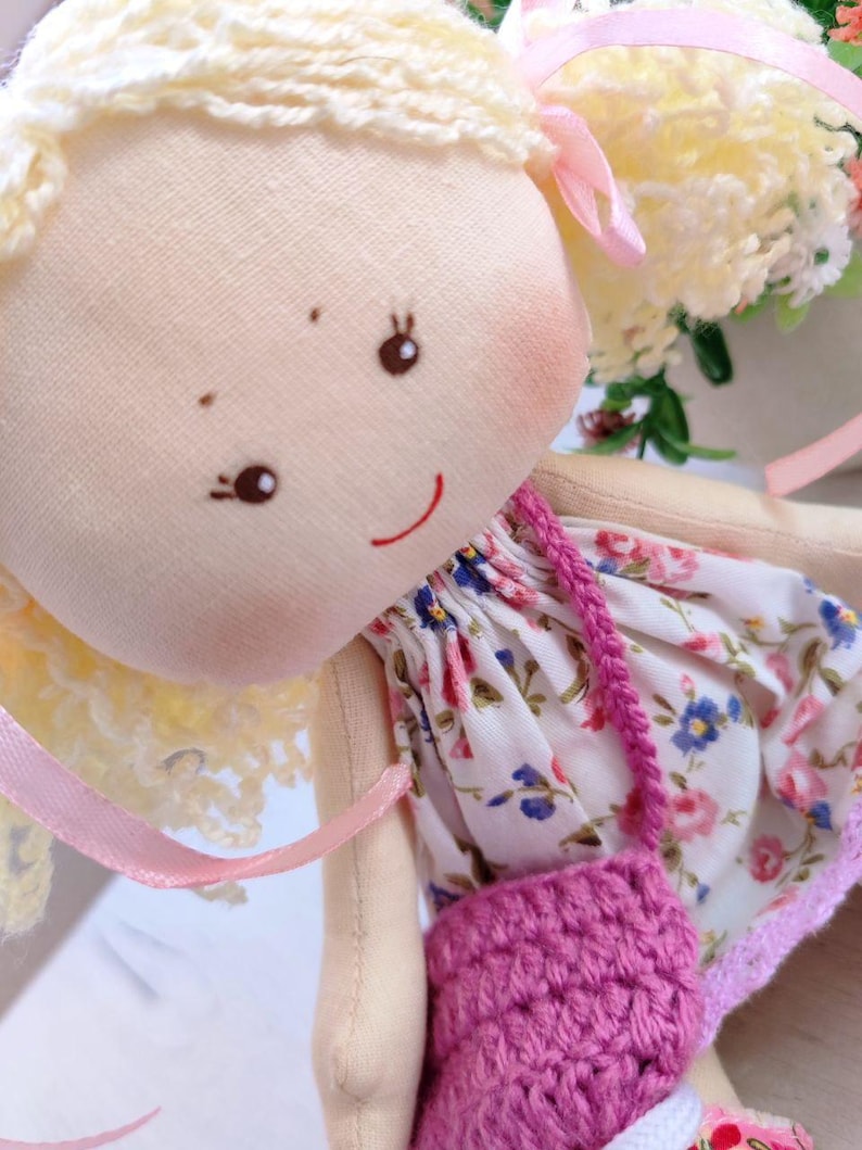 May include: A handmade cloth doll with blonde yarn hair and a painted face. The doll wears a floral print dress with a pink crocheted purse. Pink ribbons accent the doll's hair and dress, creating a whimsical and charming aesthetic.