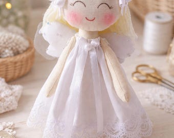 May include: A handmade angel doll with a smiling face, rosy cheeks, and blonde yarn hair tied with white bows. The doll wears a white dress with lace trim and attached white wings. The doll is approximately 8 inches tall.
