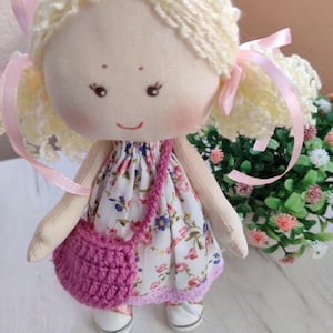 May include: A handmade cloth doll with blonde yarn hair styled in pigtails, adorned with pink ribbons. The doll wears a floral print dress, a pink crocheted purse, and white shoes. The doll's face features embroidered eyes and a smiling mouth.