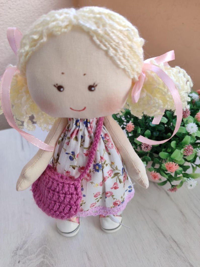 May include: A handmade cloth doll with blonde yarn hair styled in pigtails, adorned with pink ribbons. The doll wears a floral print dress, a pink crocheted purse, and white shoes. The doll's face features embroidered eyes and a smiling mouth.