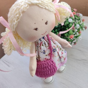 May include: A handmade doll with blonde yarn hair tied with pink ribbons. The doll wears a floral print dress, a crocheted pink purse, and white shoes. The doll has a smiling face with rosy cheeks.