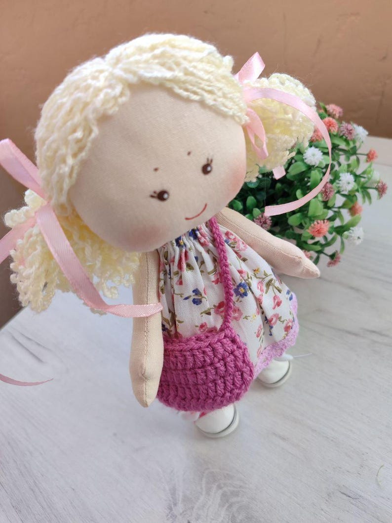 May include: A handmade doll with blonde yarn hair tied with pink ribbons. The doll wears a floral print dress, a crocheted pink purse, and white shoes. The doll has a smiling face with rosy cheeks.