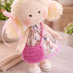 May include: A handmade cloth doll with blonde yarn hair styled in pigtails, adorned with pink ribbons. The doll wears a floral print dress, a pink crocheted purse, and white shoes. The doll has rosy cheeks and a gentle smile.