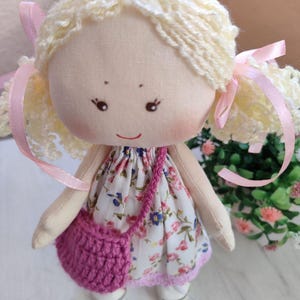 May include: A handmade cloth doll with blonde yarn hair styled in pigtails tied with pink ribbons. The doll wears a floral print dress, a crocheted pink purse, and white shoes. The doll has a painted face with rosy cheeks and a small smile.
