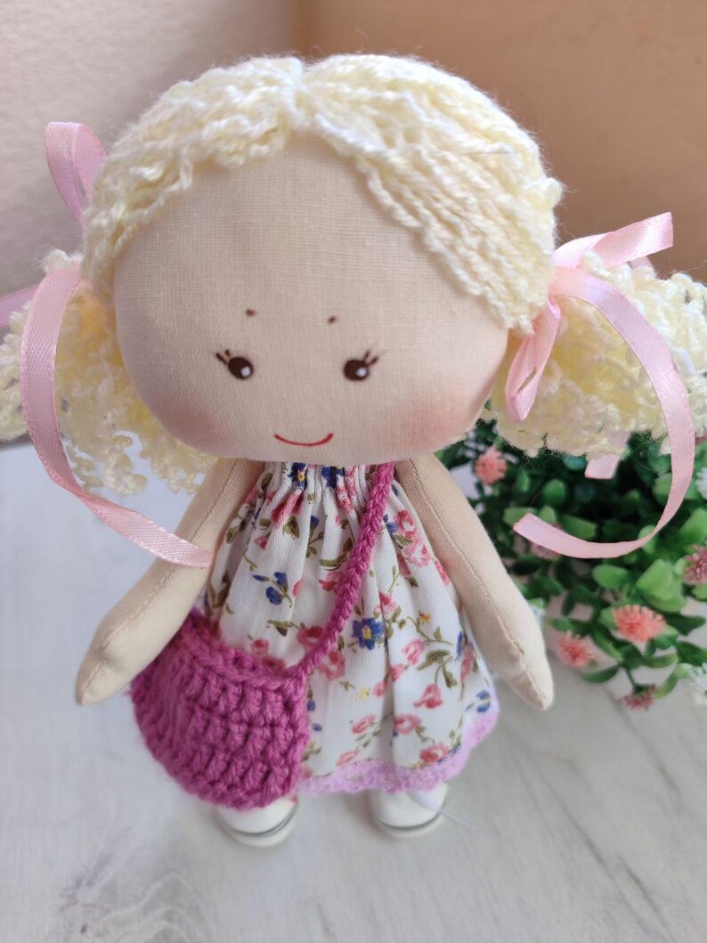 May include: A handmade cloth doll with blonde yarn hair styled in pigtails tied with pink ribbons. The doll wears a floral print dress, a crocheted pink purse, and white shoes. The doll has a painted face with rosy cheeks and a small smile.